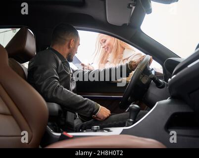 Giovane bel cliente felice uomo seduto all'interno di una nuova vettura. Bella donna sorridente venditore che chiede di firmare alcuni documenti importanti presso la concessionaria auto all'aperto. Vista dall'interno di un'auto. Foto Stock