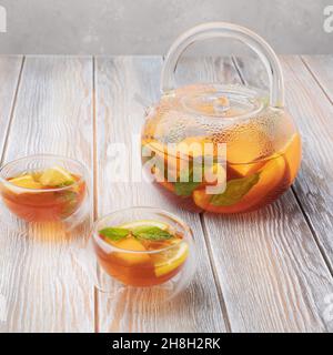 Tè di frutta con arancia, limone e menta in teiera di vetro e due tazze (ciotole) su sfondo di legno Foto Stock