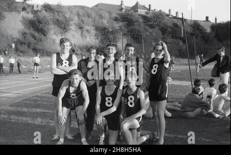 1964, storico, contea scuola sport giorno, fuori sul prato accanto a una pista di sidro, membri di una squadra di atletica della scuola, il tutto con il numero 8 sui loro giubbotti, sorridente, felice, forse esausto da una gara, posare insieme per un quadro di gruppo, Devon, Inghilterra, Regno Unito. Foto Stock