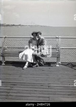 anni '50, storico, uomo e donna seduti a stretto contatto su una panchina su un molo di mare in legno. Alla fine degli anni '20 e indossando i vestiti dell'epoca, sono rilassati, tenendo le mani, magari corteggiando e/o in vacanza, Inghilterra, Regno Unito. Foto Stock