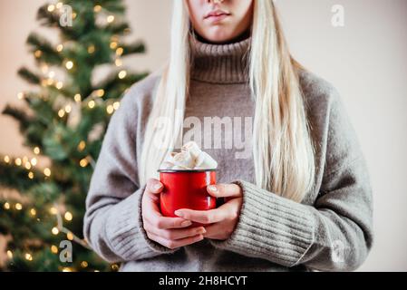 Una ragazza bionda che tiene una tazza rossa di cioccolato caldo con marshmallows su sfondo bokeh dell'albero di Natale luci Foto Stock