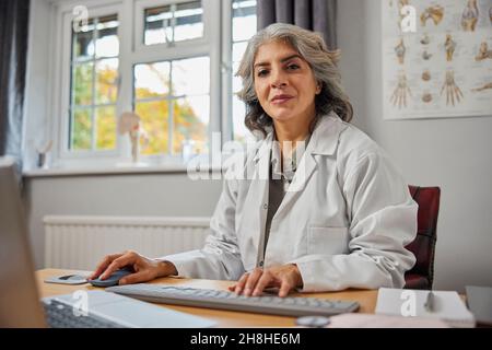 Ritratto del GP femminile maturo indossando cappotto bianco alla scrivania in ufficio medico Foto Stock
