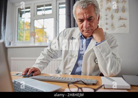 Sottoposto a stress e sopravlavorato GP maschio maturo indossando cappotto bianco alla scrivania in ufficio medici Foto Stock