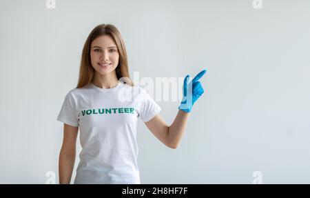 Guarda qui. Felice femmina volontario in guanti puntando da parte a spazio libero, in piedi su parete di luce e sorridendo alla macchina fotografica Foto Stock