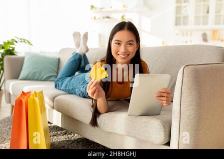 Concetto di shopping online. Signora asiatica positiva usando il tablet digitale e la carta di credito, sdraiata sul comodo divano e sorridente Foto Stock