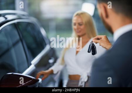 Primo piano del concessionario di auto in tuta passando le chiavi al nuovo proprietario di auto, mentre in piedi insieme al salone di auto. Sfondo sfocato di donna caucasica in piedi vicino al suo trasporto di lusso. Foto Stock