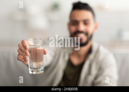 Giovane uomo arabo che mostra un bicchiere di acqua fresca e limpida alla macchina fotografica a casa, messa a fuoco selettiva. Concetto di benessere Foto Stock