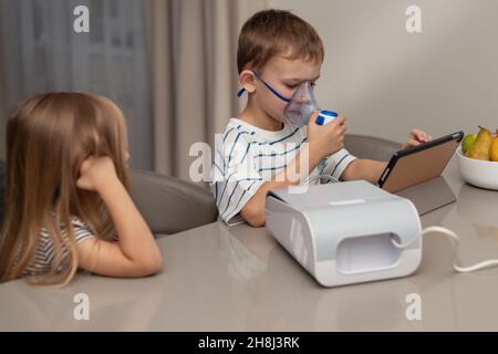 Un ragazzo in una maschera sta facendo l'inalazione, guardando un cartone animato, sua sorella è seduta vicino Foto Stock