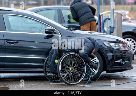 Riga, Lettonia, 4 gennaio 2021: Il conducente handicappato su sedia a rotelle pompa gli pneumatici della sua auto dal compressore in una stazione di benzina Foto Stock