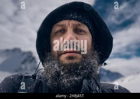 Un uomo con una barba folta, che è nel ghiaccio, guardando nella lente in montagna. È congelato nei vestiti di inverno. Foto Stock