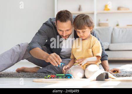 Divertimento per tutta la famiglia. Carino ragazzino che gioca ferrovia in legno e si allena con il suo papà, seduto sul pavimento di casa Foto Stock