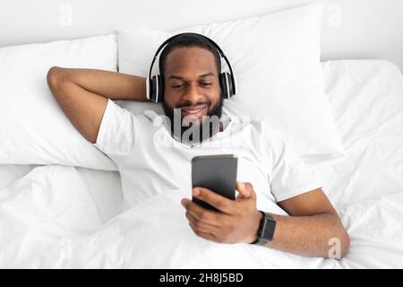 Allegro giovane afro americano barbuto ragazzo in cuffie guarda smartphone, si trova sul letto bianco Foto Stock