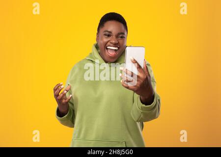 African American Lady Holding smartphone urla emotivamente su sfondo giallo Foto Stock