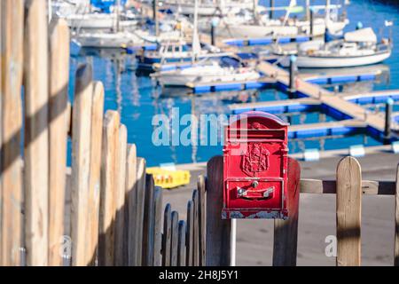 Primo piano di una cassetta postale rossa vintage montata su una recinzione di legno con un piccolo porto sullo sfondo Foto Stock