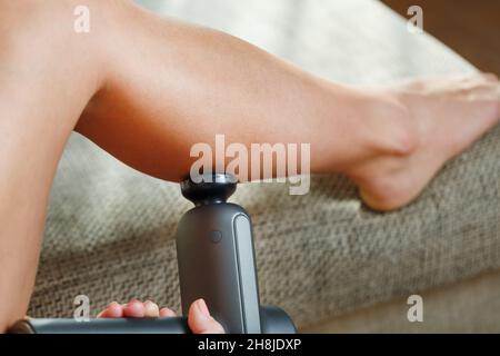 Auto-massaggio delle gambe delle donne con una pistola perfezionante a casa. Massaggio antiurto per ripristinare i muscoli della plancia e i punti di attivazione Foto Stock