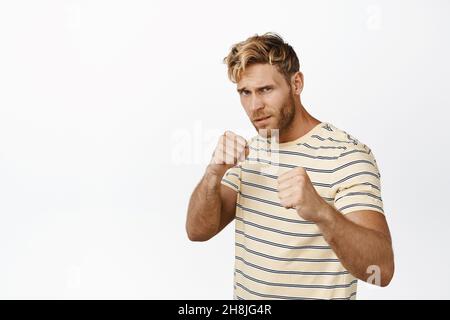 Il biondo forte e bello si trova in posizione da pugile, battendo i pugni pronti per la lotta, in posa in T-shirt casual su sfondo bianco Foto Stock