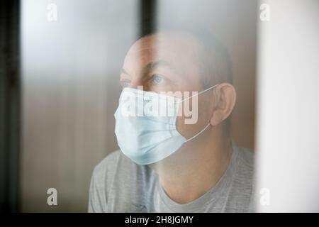 Uomo con una maschera di faccia seduta in una quarantena domestica e guardando attraverso la finestra. Foto Stock