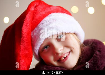 Ritratto di una ragazza felice di Natale in un cappello rosso di Santa, sorriso. Le luci si illuminano sullo sfondo. Foto Stock