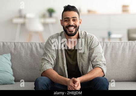 Ritratto di bel giovane arabo sorridente e guardando la macchina fotografica, seduto sul divano a casa Foto Stock