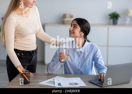 Giovane donna europea che aiuta a sorridere la signora indiana con un bicchiere d'acqua sul posto di lavoro con un computer portatile Foto Stock