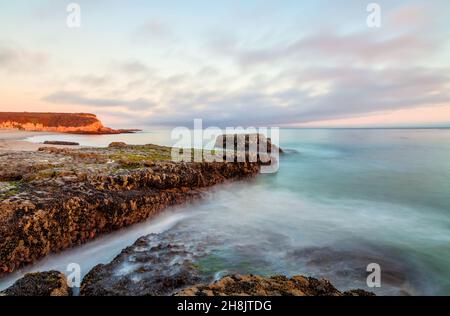 L'immagine cattura una tranquilla scena costiera al tramonto, caratterizzata da rocce scoscese ricoperte di muschio che si proiettano nell'oceano turchese e calmo. Foto Stock