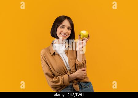 Ritratto di ragazza felice che tiene mela matura e sorride alla macchina fotografica, posando su sfondo giallo studio Foto Stock