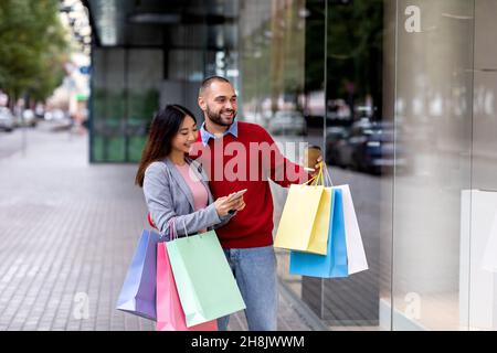 Coppia giovane e allegra con borse regalo che guardano la vetrina del negozio, controllando l'app di sconto mobile vicino al centro commerciale della città Foto Stock