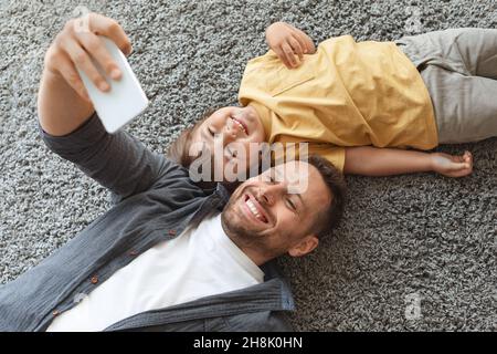 Buon ritratto di famiglia. Giovane padre che fa selfie con il figlio piccolo, sorridendo allo smartphone, riposandosi insieme a terra Foto Stock