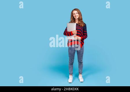 Ritratto a tutta lunghezza di donna sorridente in maglia casual a righe, in piedi tenendo chiuso il laptop o cartella, guardando la fotocamera con espressione positiva. Studio interno girato isolato su sfondo blu. Foto Stock