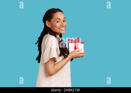 Vista laterale di donna sorridente con dreadlocks che tiene la scatola del regalo, congratulandosi, estremamente felice di ottenere il presente, guardando la macchina fotografica, indossando la camicia bianca. Studio interno girato isolato su sfondo blu. Foto Stock