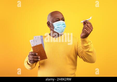 Ritratto di turista maschile nero senior in maschera di protezione con passaporto, biglietti e aereo giocattolo su sfondo arancione studio Foto Stock