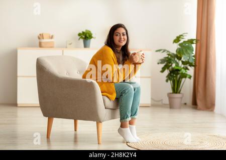 Tutta la lunghezza di positiva signora araba seduta in poltrona con tazza di caffè caldo e sorridente alla macchina fotografica, spazio copia Foto Stock