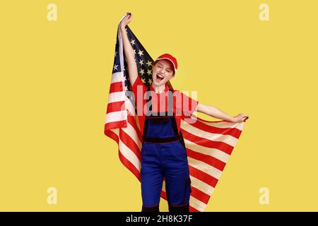 Donna operaia eccitata in piedi con la bandiera americana nelle mani e urlando felicemente, festeggiando la festa, indossando l'uniforme di lavoro e il cappuccio rosso. Studio interno girato isolato su sfondo giallo. Foto Stock