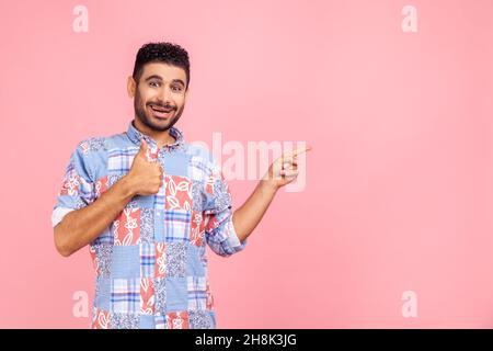 Uomo felice sorridente con barba che indossa una maglietta casual blu rivolta da parte e con il pollice rivolto verso l'alto, presentando spazio per il testo promozionale. Studio interno girato isolato su sfondo rosa. Foto Stock