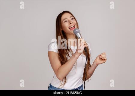 Ritratto di bella donna positiva cantare forte canzone, tenere il microfono in mano, divertirsi riposando in karaoke, indossare T-shirt bianca. Studio interno girato isolato su sfondo grigio. Foto Stock