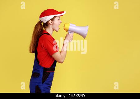 Ritratto di vista laterale di donna lavoratore arrabbiata urlando forte in megafono, dando il comando ai suoi costruttori subordinati, indossando uniforme da lavoro e berretto rosso. Studio interno girato isolato su sfondo giallo. Foto Stock