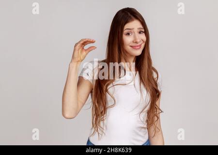 Ritratto di donna infelice con capelli scuri che mostrano piccoli gesti di dimensioni con espressione del viso delusa e frustrata, indossando una T-shirt bianca. Studio interno girato isolato su sfondo grigio. Foto Stock