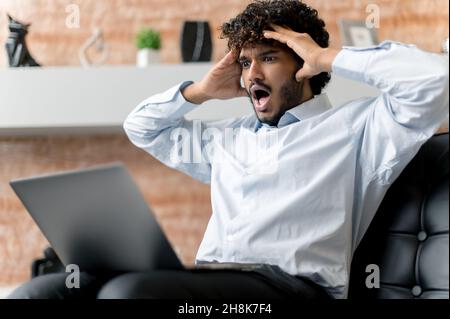 Eccitato stupito, indignato uomo indiano, uomo d'affari o freelance, seduto su una sedia, guardando un computer portatile in sorpresa, tenendo la testa con le mani, vedendo notizie inaspettate Foto Stock