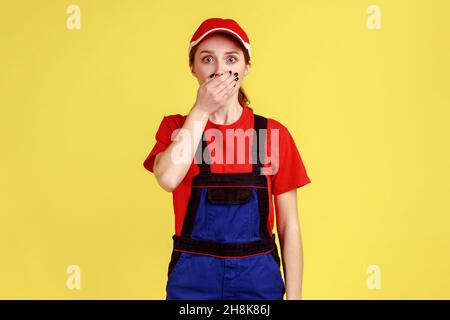 Ritratto della donna operaia stupita scioccata in piedi e guardando la macchina fotografica con gli occhi grandi, coprendo la bocca con il palmo, indossando uniforme da lavoro e cappuccio rosso. Studio interno girato isolato su sfondo giallo. Foto Stock