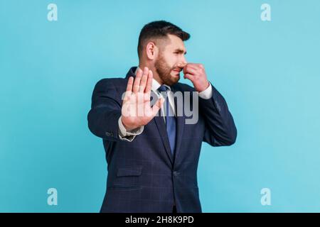 Odore sgradevole. Giovane uomo adulto bearded che indossa il vestito ufficiale di stile che tiene il respiro con le dita sul naso, disgustato da cattivi odori, arresto gesturante. Studio interno girato isolato su sfondo blu. Foto Stock