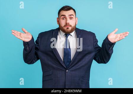 L'uomo senza indizio perplesso con la barba che indossa abiti di stile ufficiale che si stendono le mani e le spalle, non sicuro e indifferente in alcune domande. Studio interno girato isolato su sfondo blu. Foto Stock