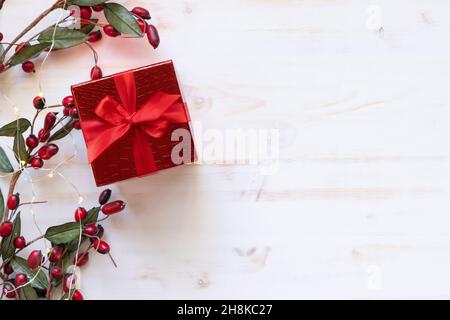 Un regalo di Natale con arco a nastro rosso dall'alto con un bordo di acini rossi e mini luci natalizie su sfondo bianco con spazio copia Foto Stock