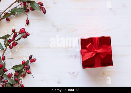 Un regalo di Natale con arco a nastro rosso dall'alto con un bordo di acini rossi e mini luci natalizie su sfondo bianco con spazio copia Foto Stock