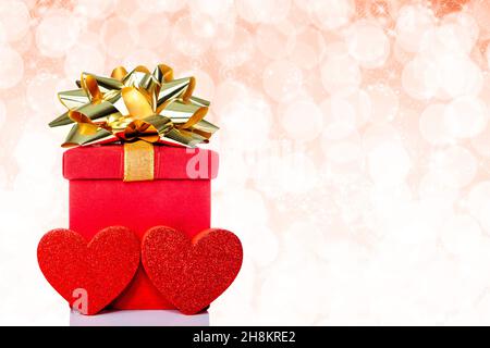 Scatola regalo rossa con arco dorato e cuori su sfondo scintillante con spazio copia. Adatto per feste come Natale, matrimonio, San Valentino, o Foto Stock