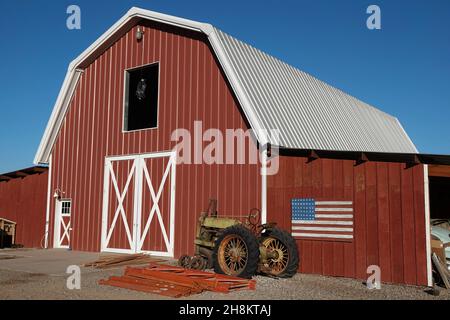 Un vecchio trattore verde arrugginito di fronte ad un granaio rosso con una bandiera americana di legno fatta in casa sul muro. Foto Stock