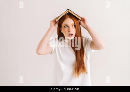 Ritratto dello studio di giovane studentessa stressata che copre la testa con libro che guarda via, in piedi su sfondo bianco isolato. Foto Stock