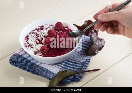 Una mano della donna tiene un cucchiaio sopra una ciotola di frullati fatti di mele di barbabietola e lamponi. Cibo vegano a base di piante Foto Stock