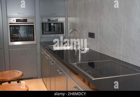 Un moderno design interno della cucina con un piano cottura nero con un lavello e un piano cottura elettrico, armadi grigi, un forno incorporato e una macchina per il caffè. Foto Stock