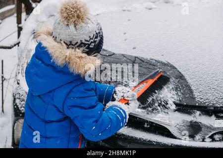 Il ragazzo piccolo carino pulisce la neve fuori dell'automobile al cortile d'inverno. Bambino piccolo ragazzo del toddler che aiuta il padre a liberare l'automobile della neve ed usando lo strumento della spazzola per Foto Stock