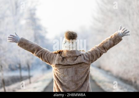 Donna che indossa un cappotto in pelliccia e un cappello in maglia sta godendo l'inverno all'aperto. Vista posteriore della donna con le braccia sollevate. Clima freddo e alberi ghiacciati nel parco Foto Stock